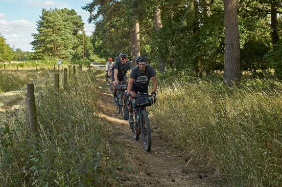 Cyclists riding along a sandy path by the edge of a wood.